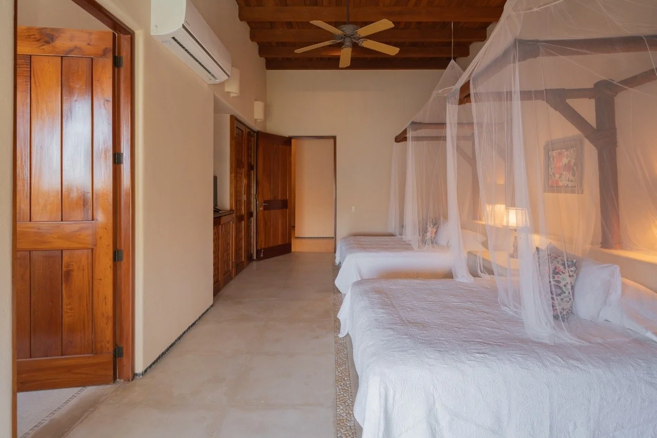 Habitación con dos camas, mosquiteros, paredes blancas, techos de madera, puertas de madera y aire acondicionado.