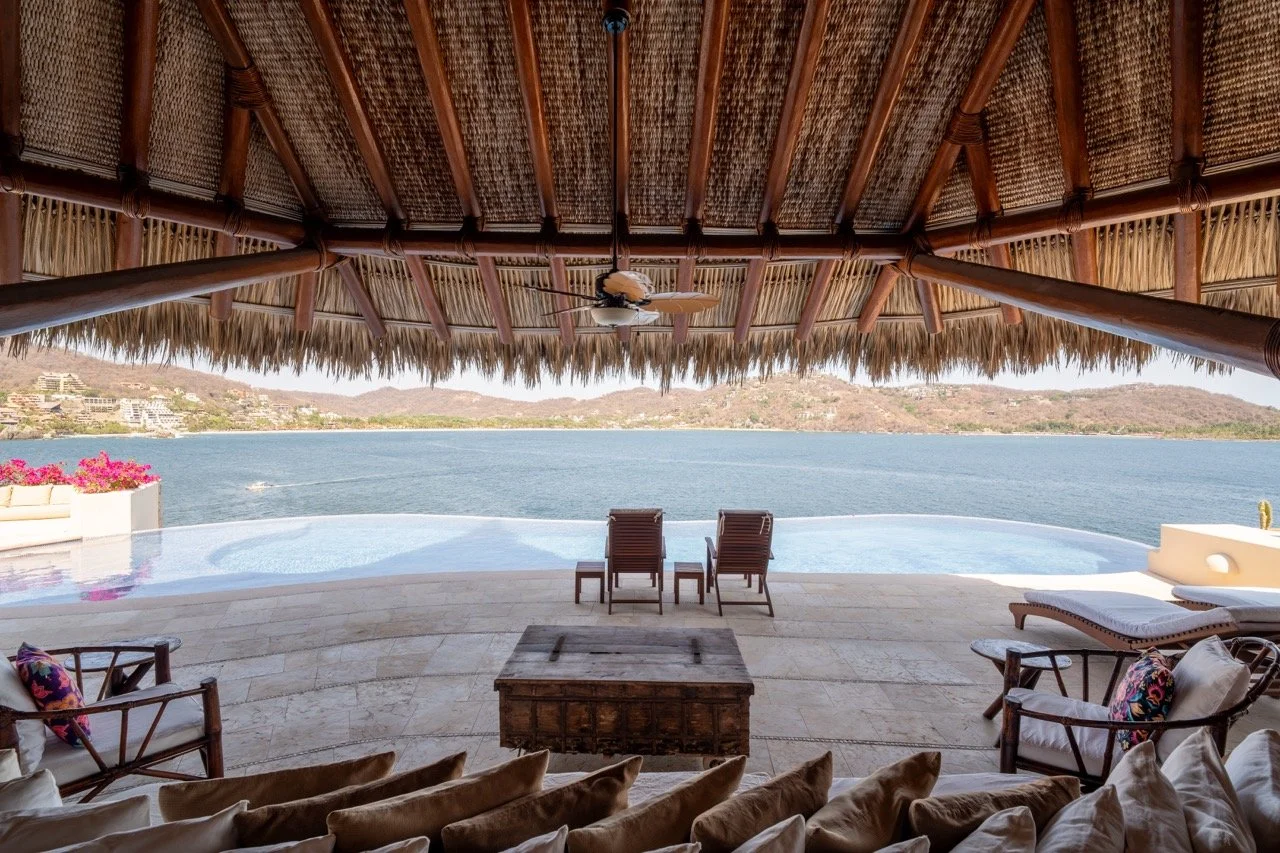 Vista de una piscina infinita con vista al mar desde una terraza con sillones y sombrilla de palma quechua.