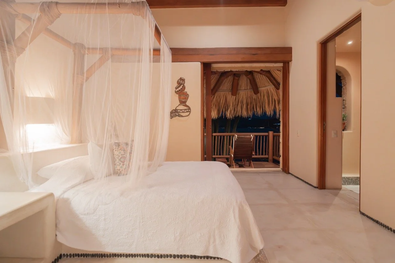 Habitación con cama con dosel, cortinas de malla, y balcón con vista nocturna, decoración en estilo tropical con madera y paja.
