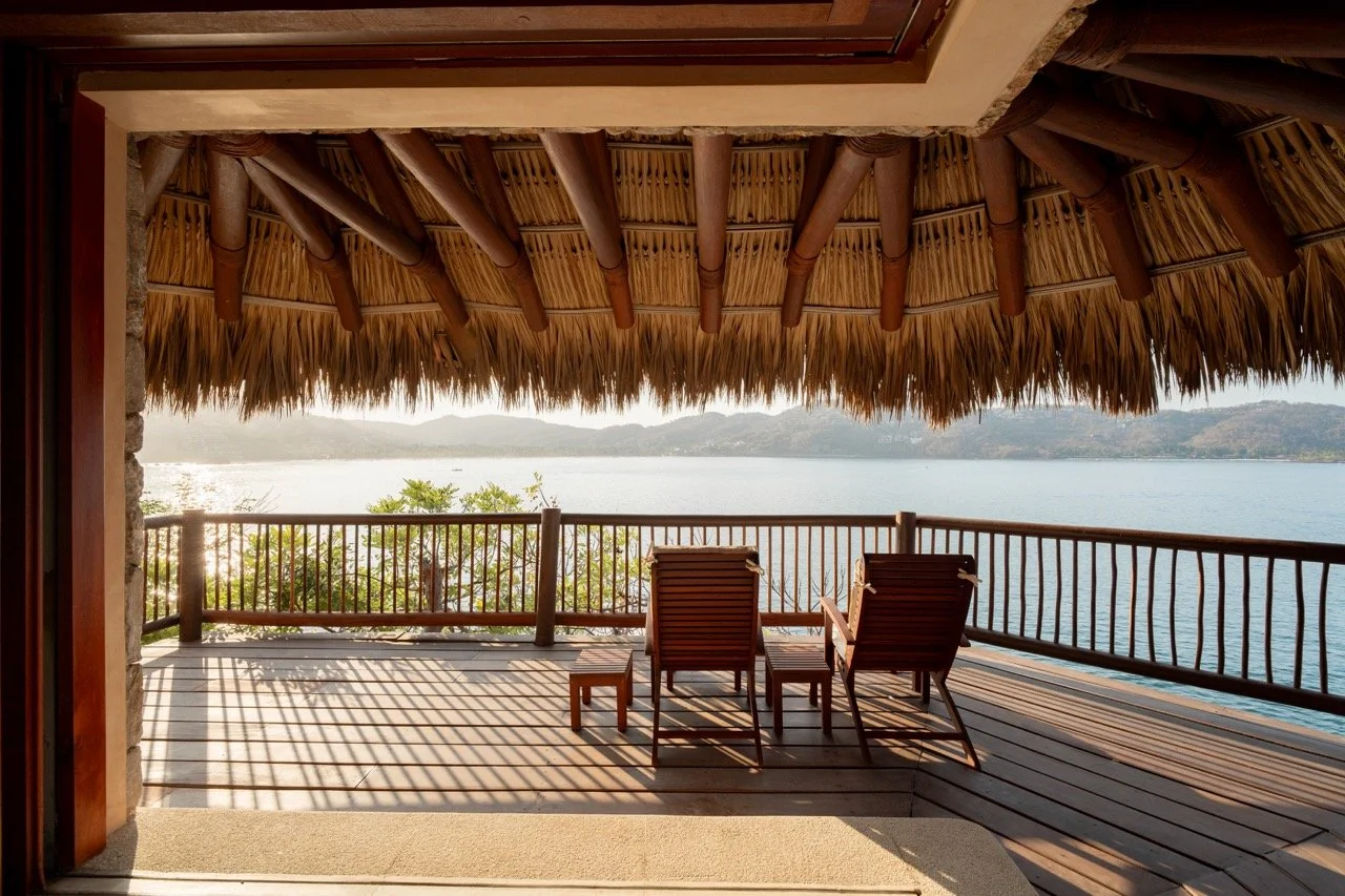 Vista desde una terraza de madera con sillas y mesa de madera, techo de palma y vista a un lago con montañas al fondo.