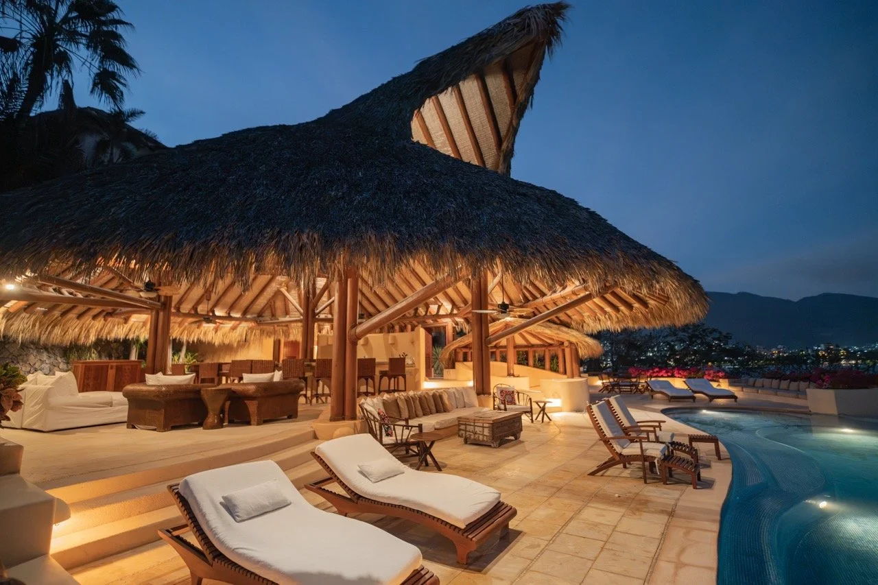 Zona de alberca con sillones y sombrillas bajo una palapa en un atardecer tropical.