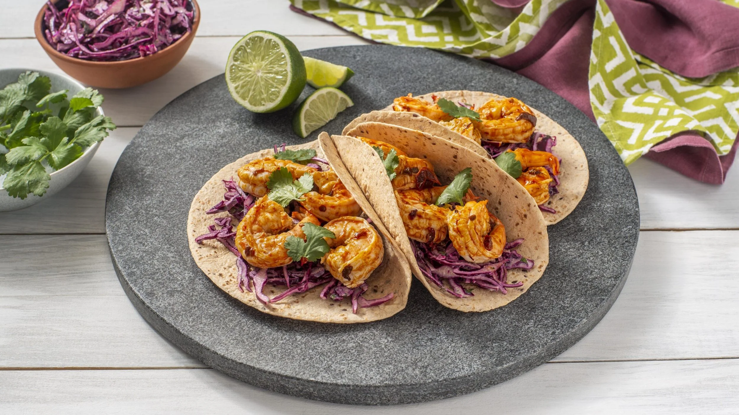 Tacos de camarón sobre plato con repollo morado, cilantro y limones en un fondo de mesa de madera. Bastante colorido y apetitoso.