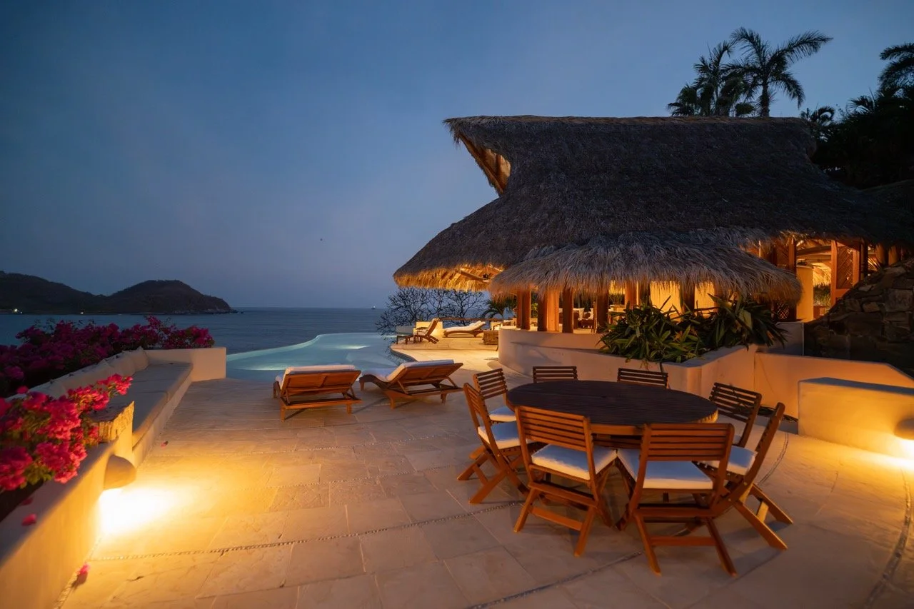 Una terraza de noche con vista al mar, mobiliario de madera y un techo de paja en un ambiente tropical, decorado con plantas y flores coloridas.