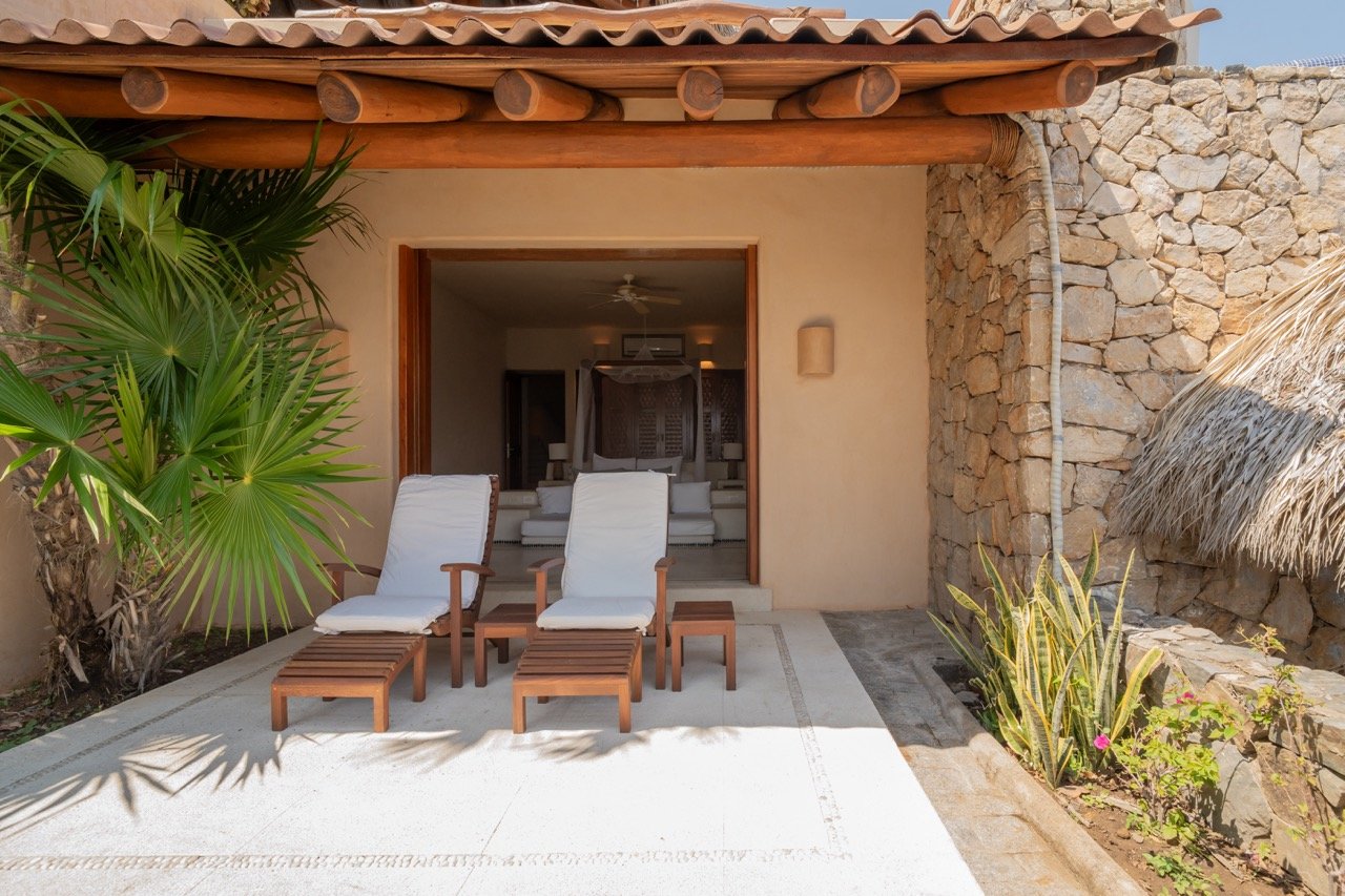 Vista de una terraza en un hotel o casa de playa con dos sillones reclinables de madera y cojines blancos, plantas tropicales y pared de piedra