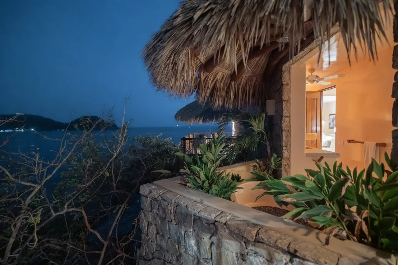 Vista nocturna desde una terraza de un hotel o cabaña junto al mar con paredes de piedra y techo de palma, mostrando plantas verdes y la distancia del mar con islas en el horizonte.