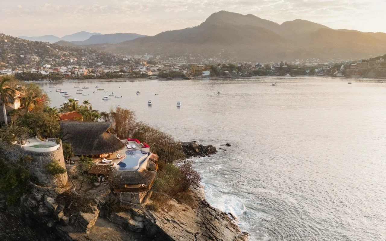 Vista del río con botes, junto a casas y un hotel con piscina en la orilla, con montañas al fondo durante el atardecer.