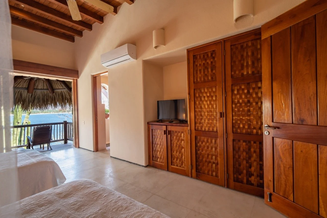 Habitación con puertas de madera, televisor en la esquina y vista a un balcón con vista al mar y palapas.