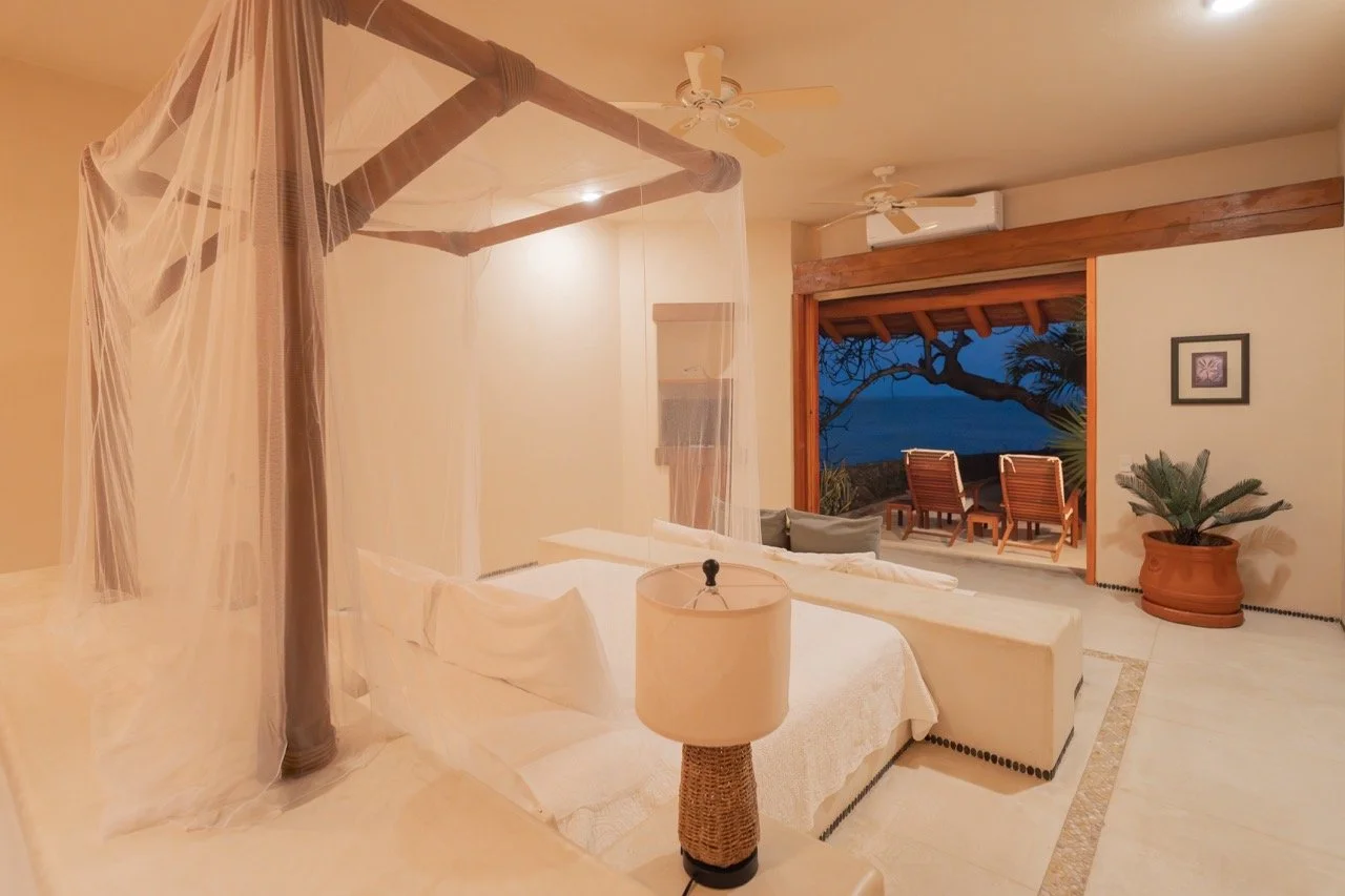 Habitación con cama con mosquitero, ventana abierta a una vista de mar con sillas y árboles, planta en maceta, lámpara y ventiladores en el techo.