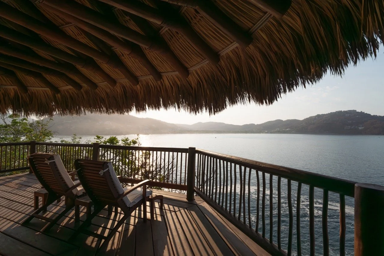 Vista desde una palapa de madera y techo de palma, con dos sillas junto al balcón y vista a un cuerpo de agua, con colinas en el fondo.
