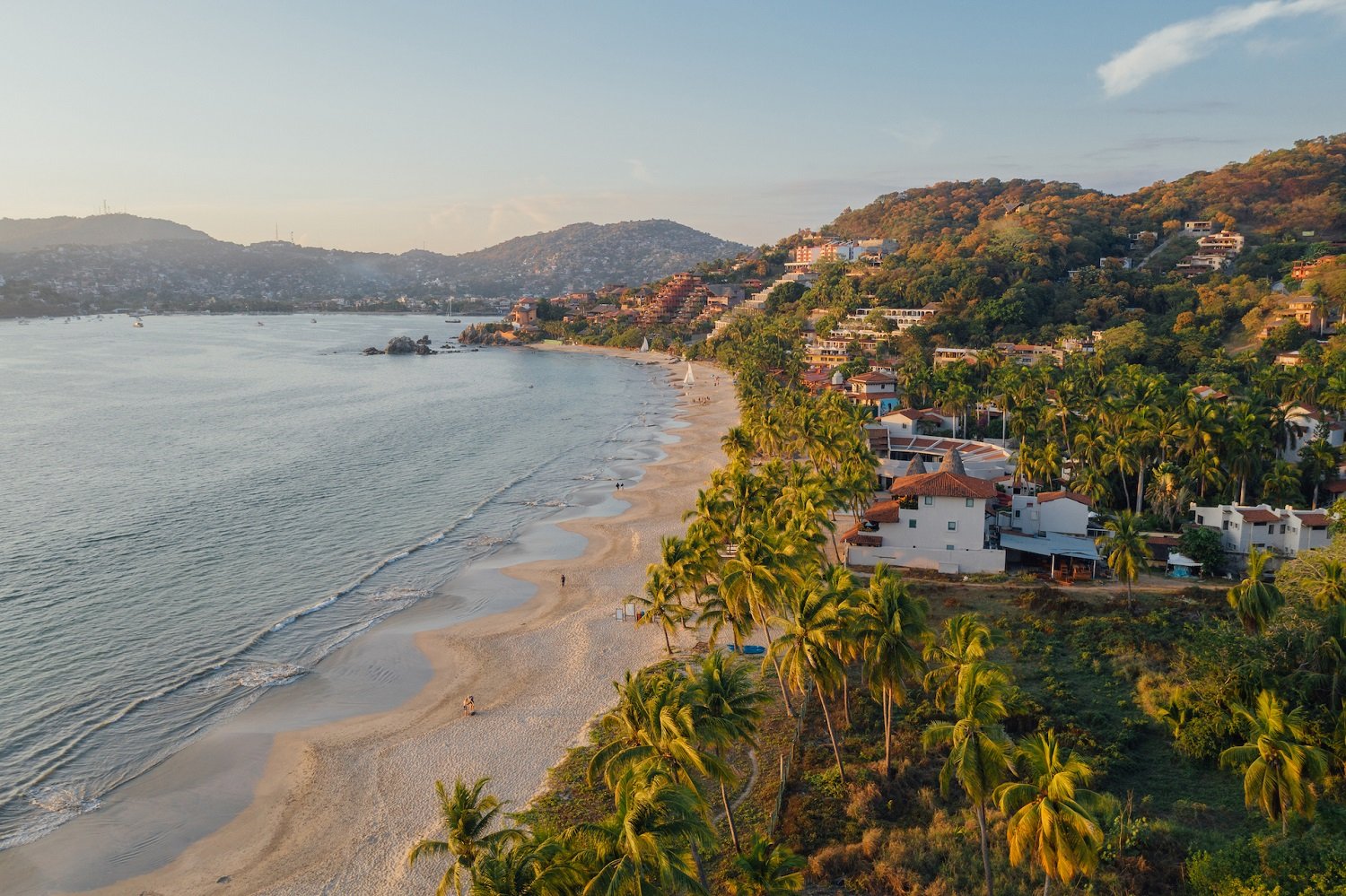 Vista aérea de una playa con arena clara, aguas tranquilas y palmeras, rodeada de casas y colinas verdes.