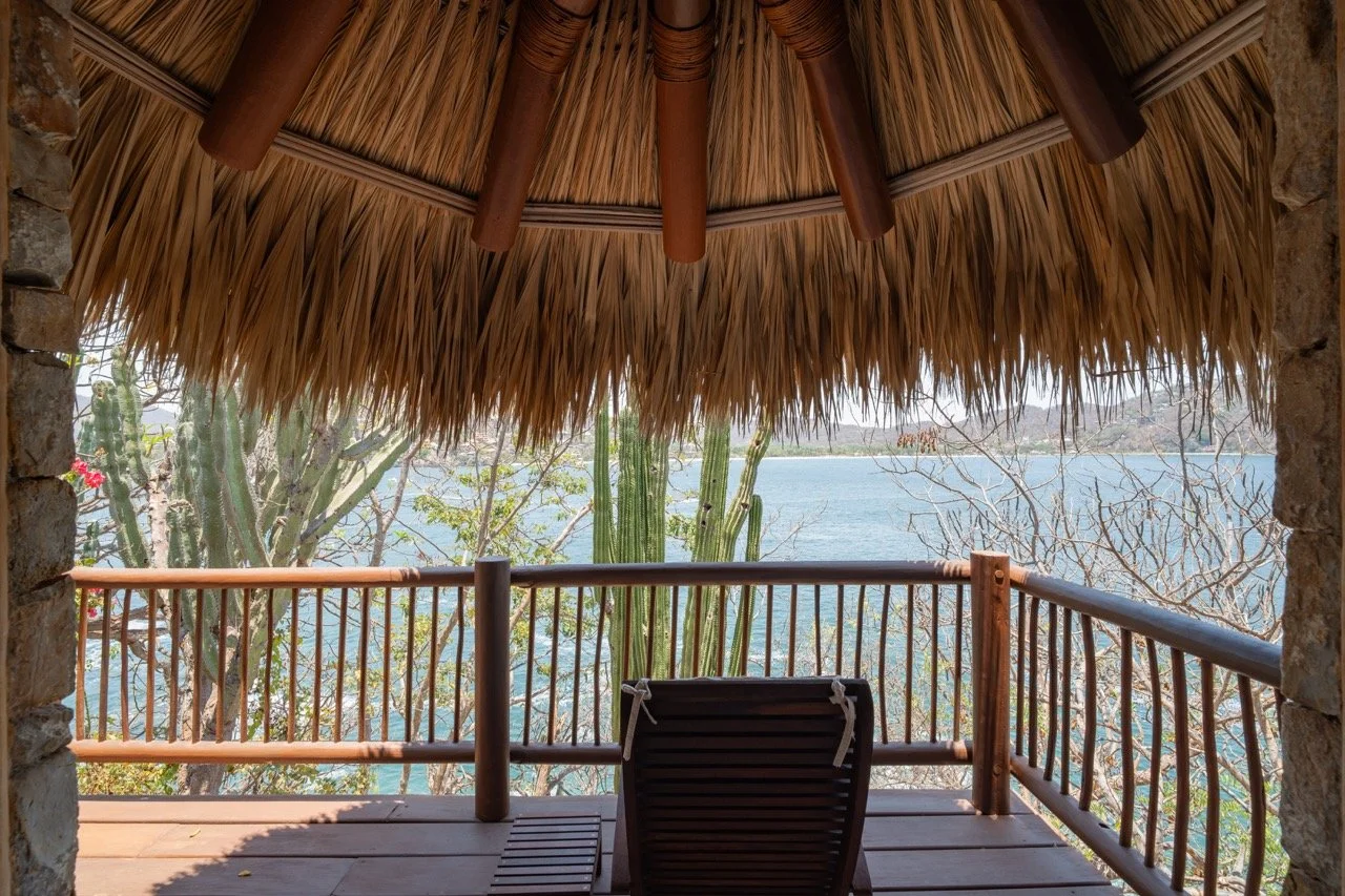 Vista desde una terraza con una silla de madera, techo de hojas de palma y vista a un lago rodeado de árboles y cactus.