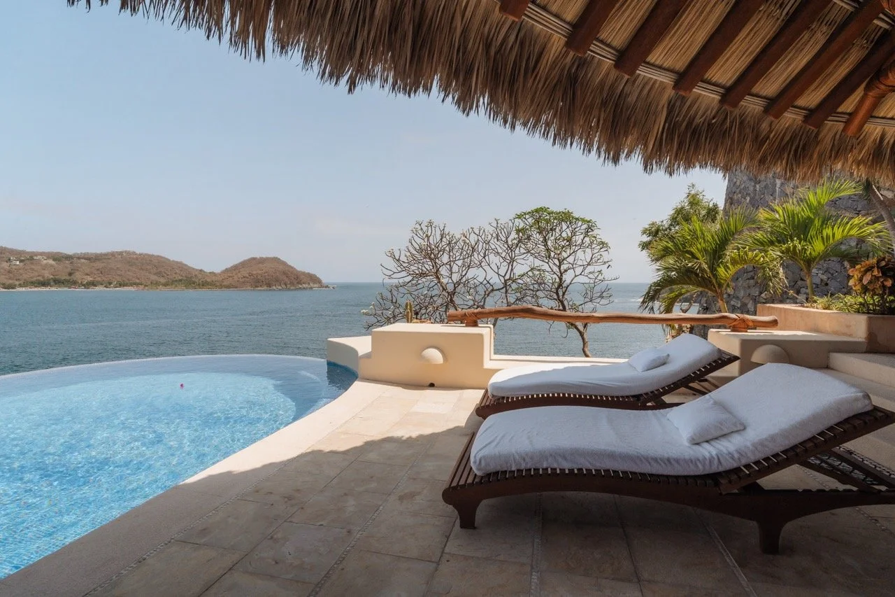 Vista de una terraza con dos tumbonas de mimbre y cojines blancos, una piscina pequeña con vista al mar y una zona con plantas y árboles, bajo un techo de hojas de palmera.