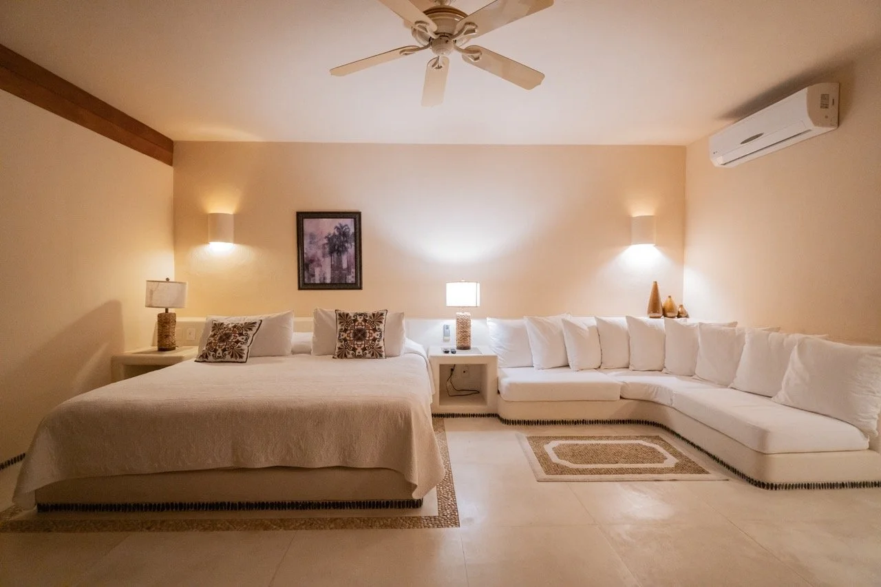 Habitación con cama grande, sofá blanco en esquina, lámparas, cuadro en la pared y aire acondicionado.