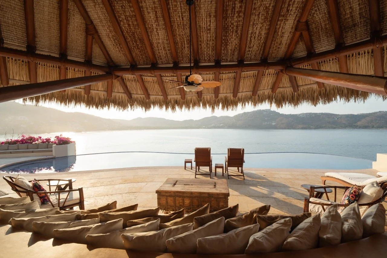 Vista de una terraza con sillas, tumbonas y un toldo de paja, con vista a un lago y montañas al atardecer.