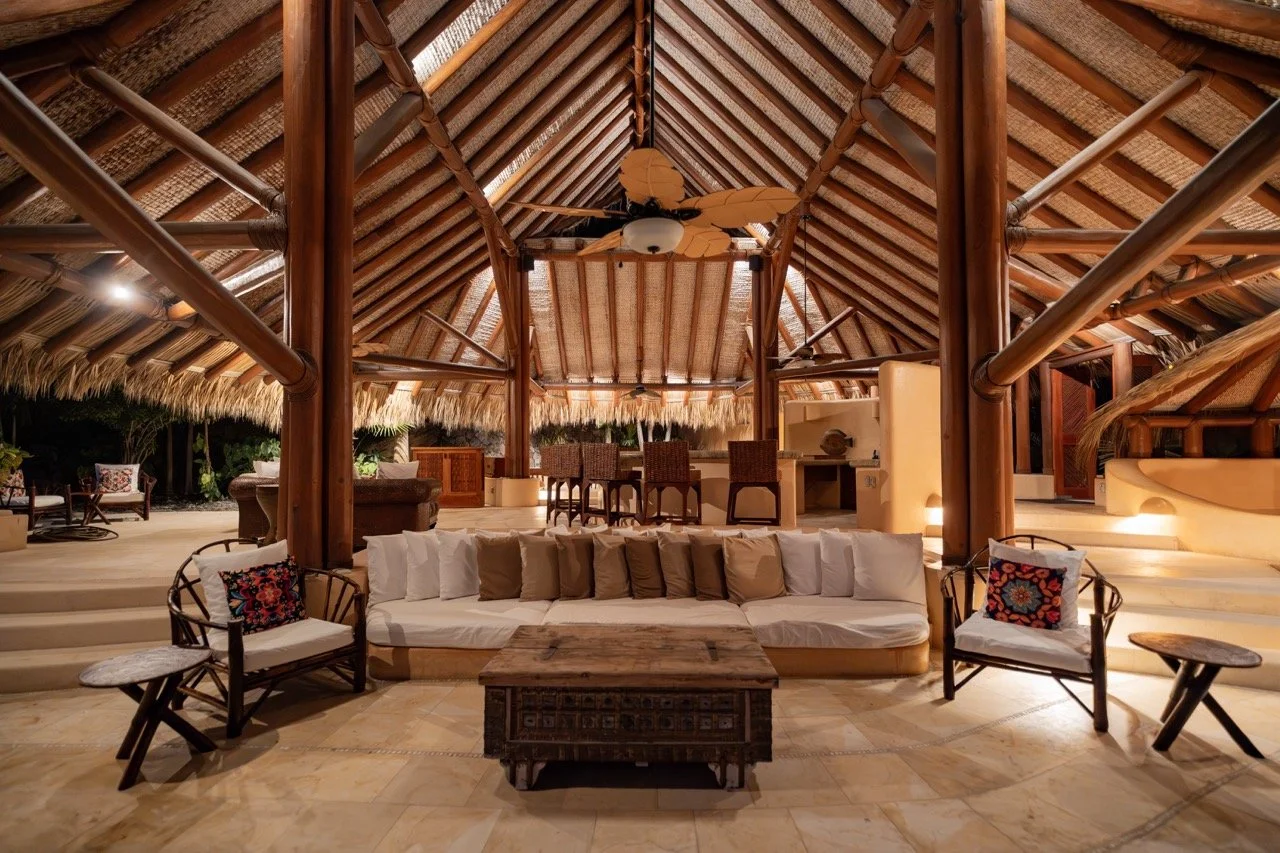 Interior de casa de madera con techo de palapa, sillones y mesas de madera, bar y área de descanso decorada con cojines, en un ambiente amplio y con iluminación cálida.