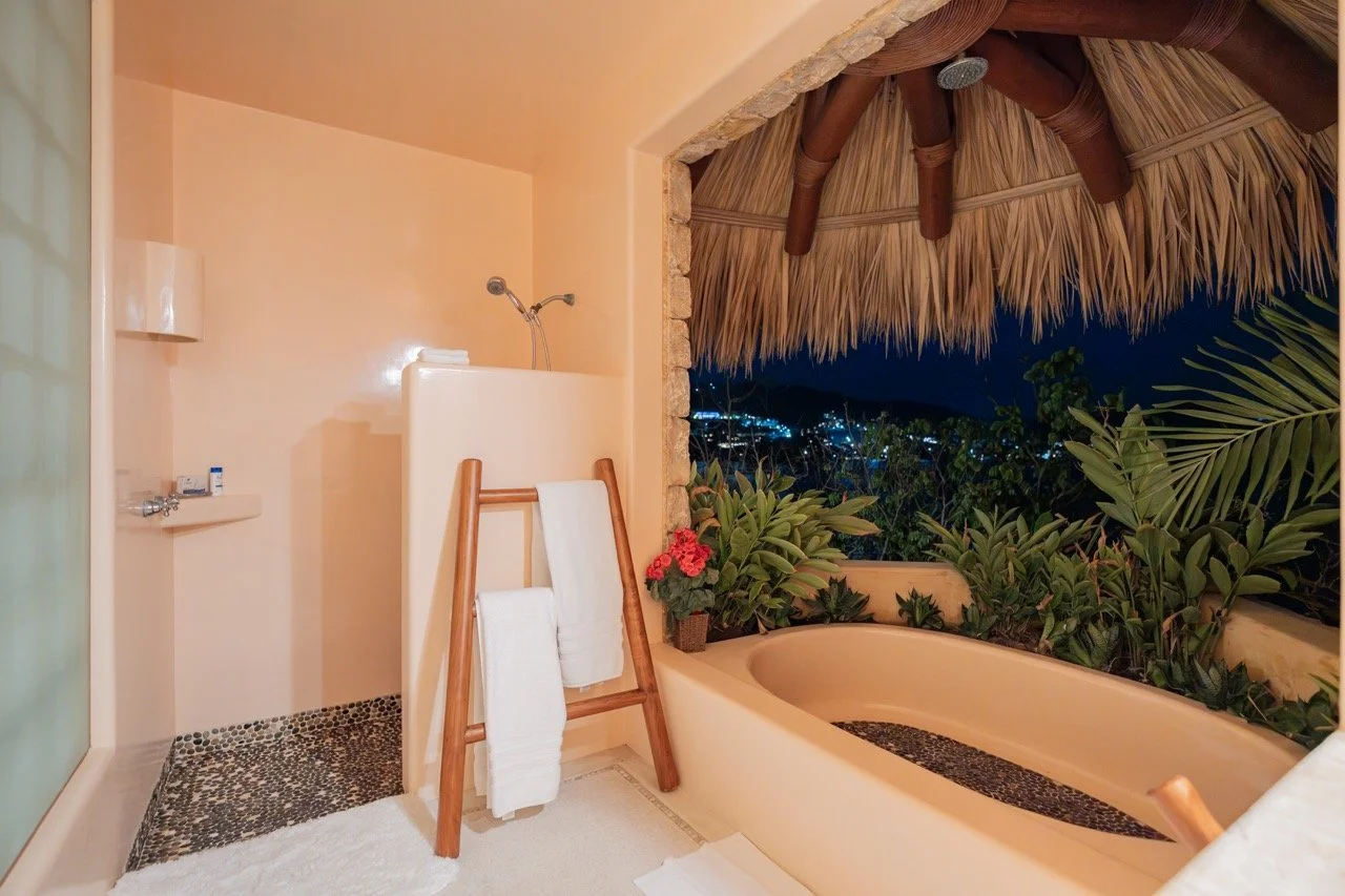 Baño con bañera al aire libre rodeada de plantas, con un techo de palma y vista nocturna hacia la ciudad.