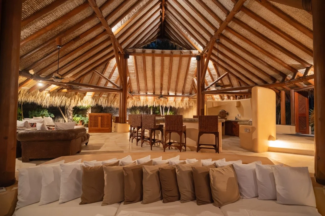 Interior de una cabaña con techos de palma, zona de bar y sala con muchos cojines blancos y beige.