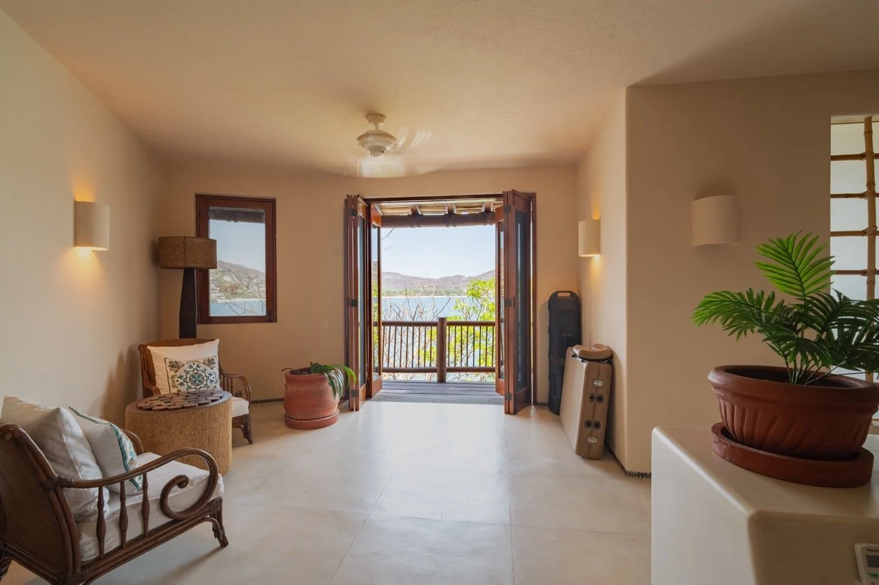 Sala con puertas abiertas a un balcón con vista a montañas y agua, muebles de madera, plantas y decoración en tonos neutros.