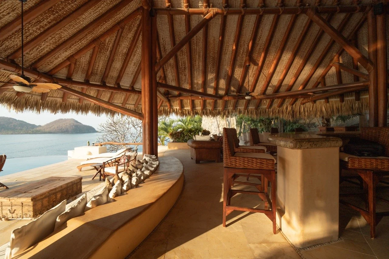 Vista de una palapa con techo de palma, vista al mar, con sillones y barra de madera y piedra, decorada con plantas y vista a una isla en el fondo.