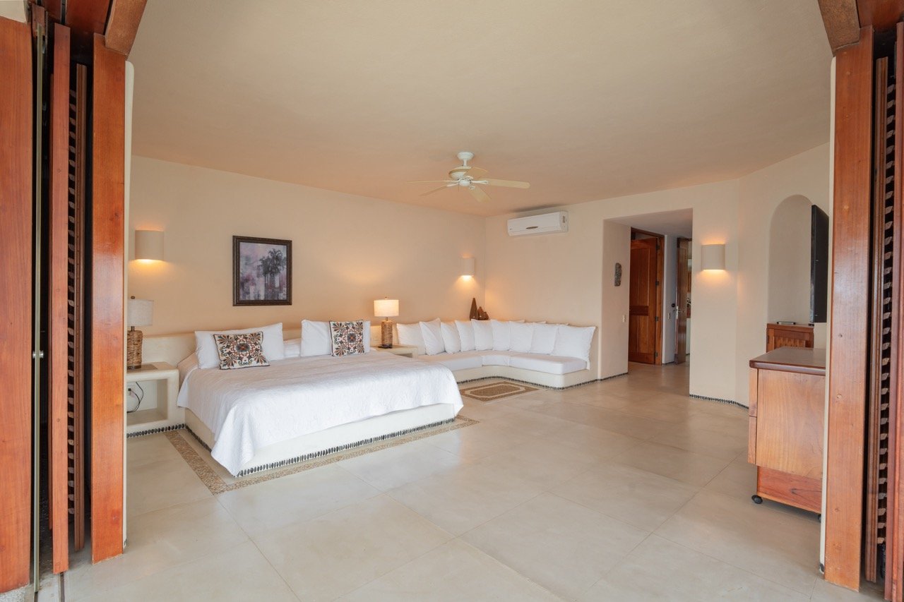 Sala con sofá blanco en forma de L, lámparas de mesa y pared, ventilador en el techo, aire acondicionado, decoraciones de pared y muebles de madera.