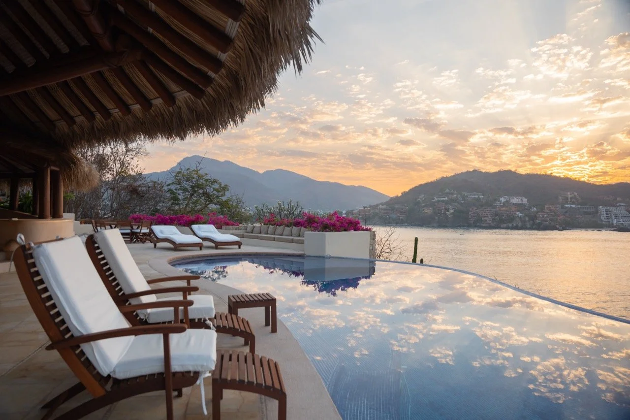 Vista de una piscina infinita con sillones de madera con cojines blancos, flores rosadas y un atardecer en un paisaje montañoso junto a un lago.