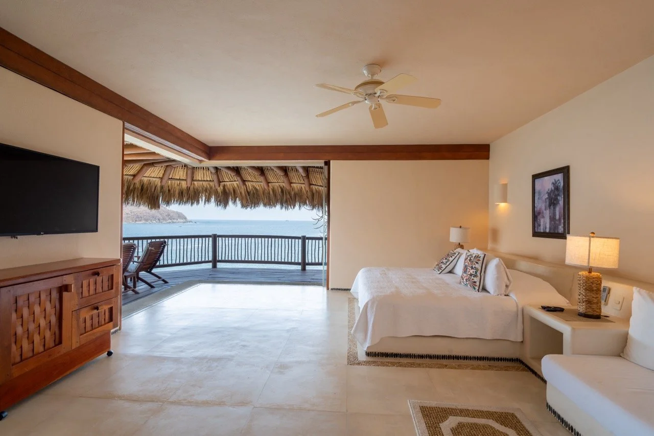 Habitación de hotel con vista al mar, cama grande con almohadas decorativas, mesa de noche con lámpara, televisión, terraza con sillones y vista a la playa