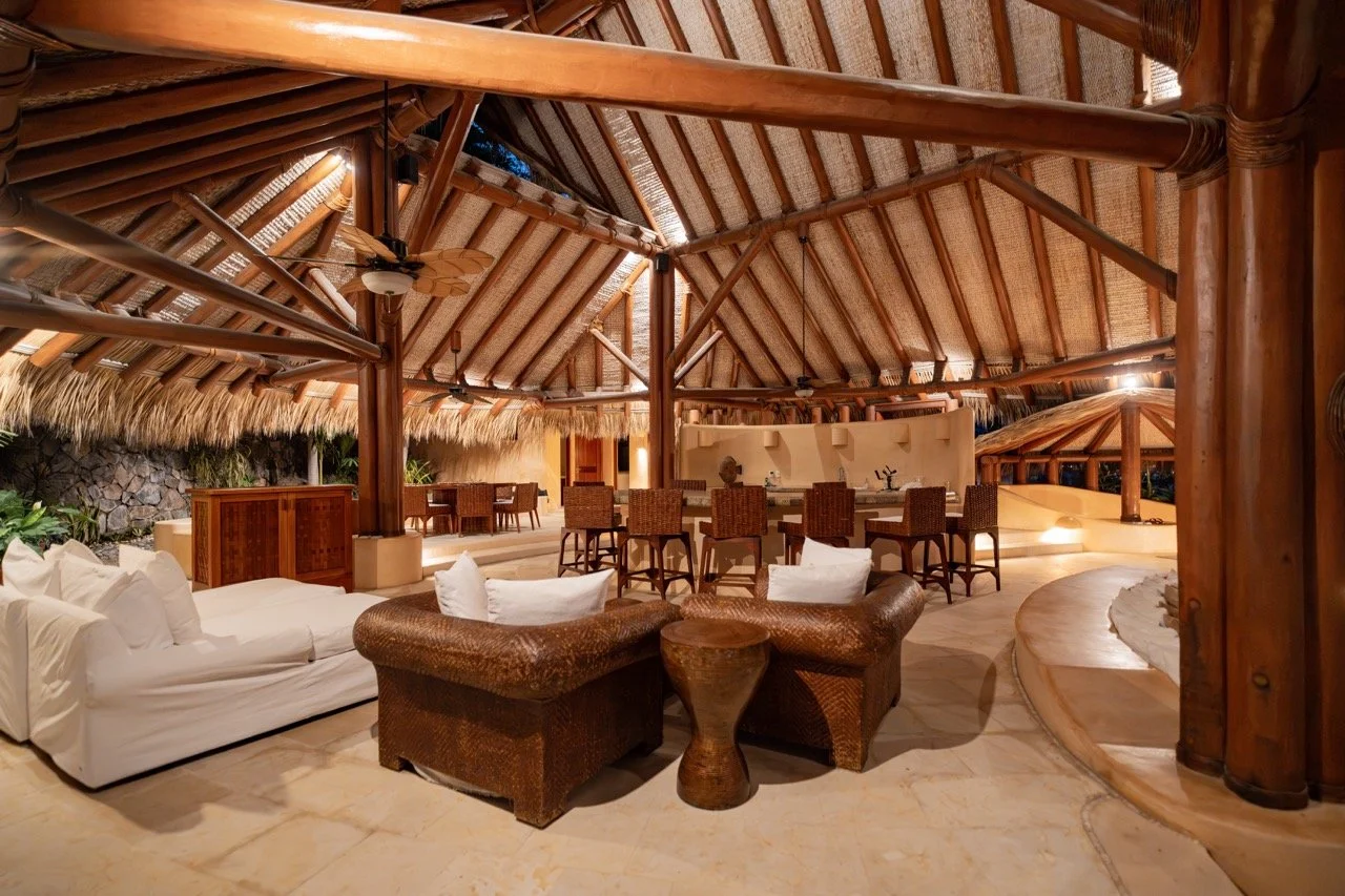 Interior de un alojamiento con estructura de madera y decoración natural, incluyendo sofás blancos, sillas de mimbre, un bar y áreas de descanso.