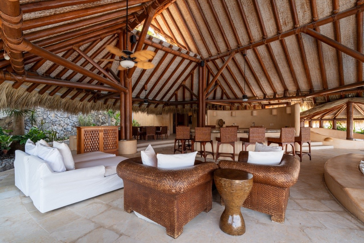Área de descanso cubierta con muebles de mimbre y sofás blancos, bar con sillas de mimbre, estructura de madera y techo de palma.