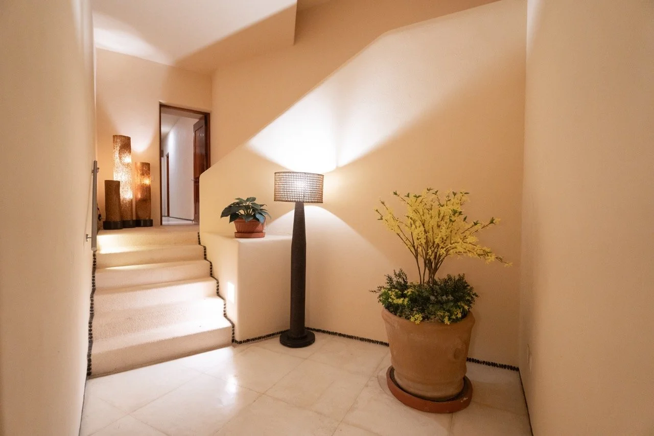 Vestíbulo interior con escaleras, una lámpara de pie, dos plantas en macetas, y decoración cálida con luz suave.