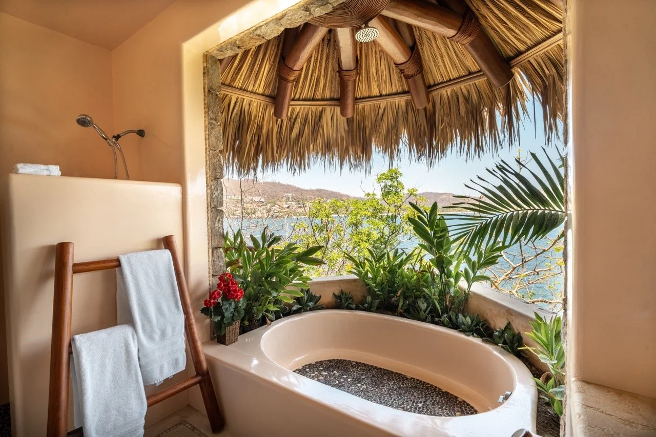 Tina de baño en una habitación con vista al mar, rodeada de plantas verdes y un techo de hojas de palma, con una estructura de madera para colgar las toallas.