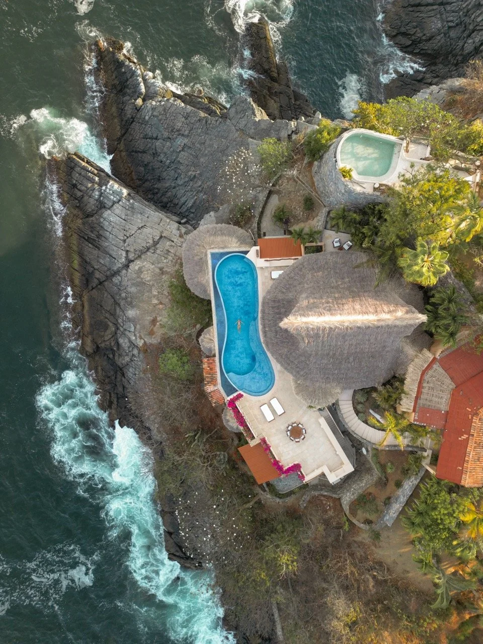 Vista aérea de una vivienda en la playa con piscina, jacuzzi y áreas verdes, rodeada de rocas y el océano.