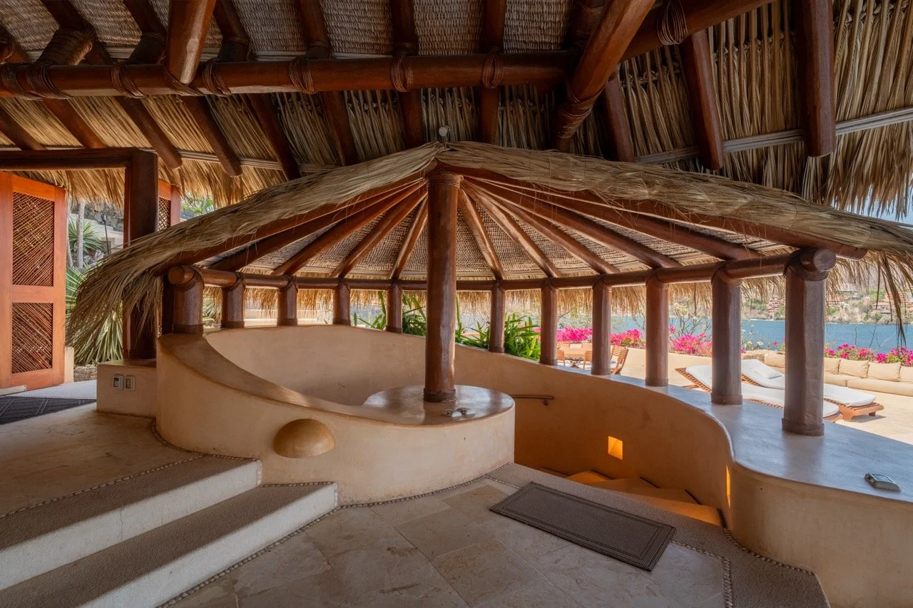 Terraza con estructura de madera y techo de palma, con una piscina en forma de cruz y vista al mar, decorada con flores y muebles de exteriores.