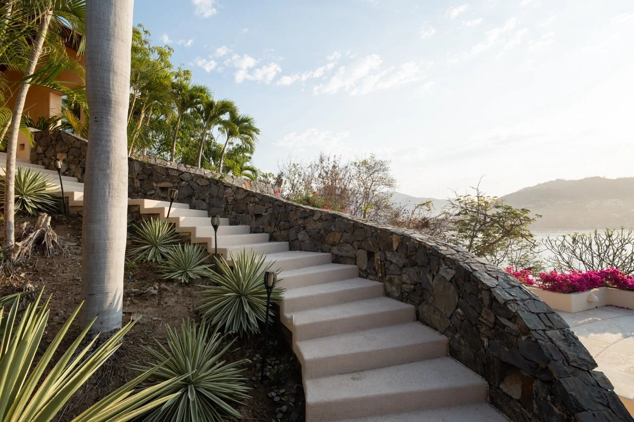 Escalera de piedra blanca con barandal de roca, rodeada de plantas tropicales y Palmeras, con vista al mar y montañas al fondo en un día soleado.