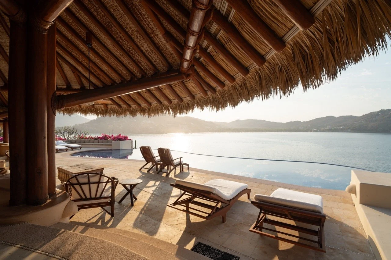 Una vista al atardecer desde una terraza junto a un cuerpo de agua, con sillas y tumbonas de madera y un borde de inflable en la piscina, bajo un techo de palma.