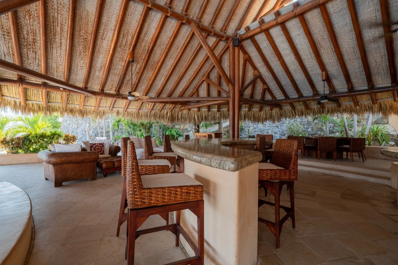 Área de descanso con mostrador de bar de madera y madera en el techo, rodeada de plantas y muebles de ratán.