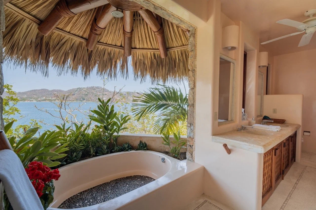 Vista de una bañera redonda en un balcón con plantas y vista al lago y montañas al fondo, espacio interior con lavabo y espejo, techo con ventilador y accesorios decorativos en tonos calidos.