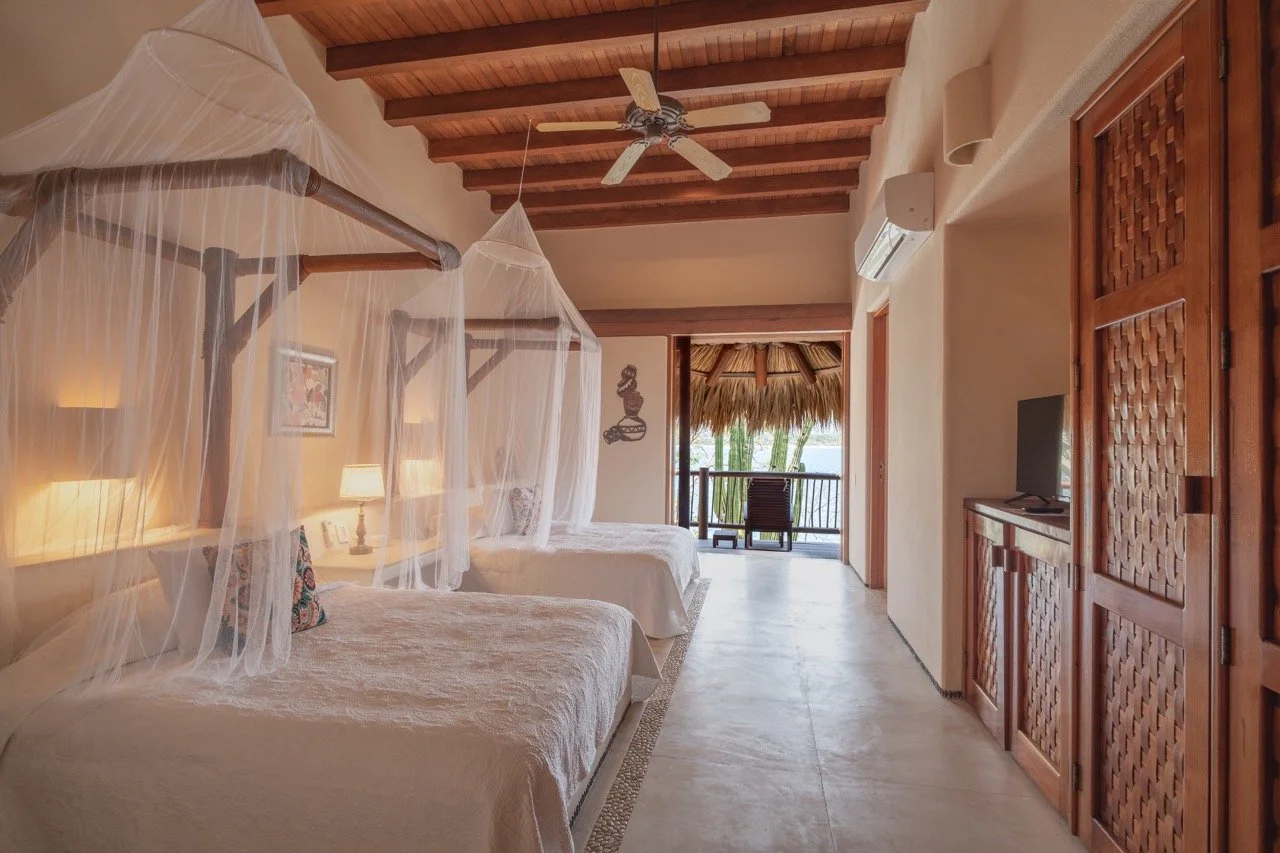 Habitación de hotel con dos camas, ventilador en el techo, balcón con vista a la naturaleza.