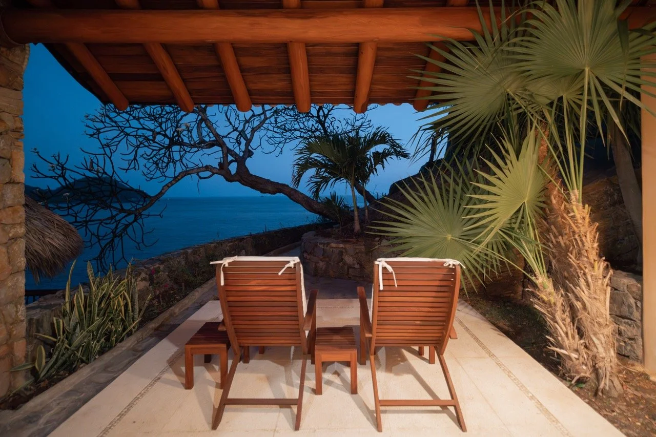 Dos sillones de madera con cojines blancos en una terraza con vista al mar, rodeada de plantas tropicales y un árbol sin hojas, en la noche.