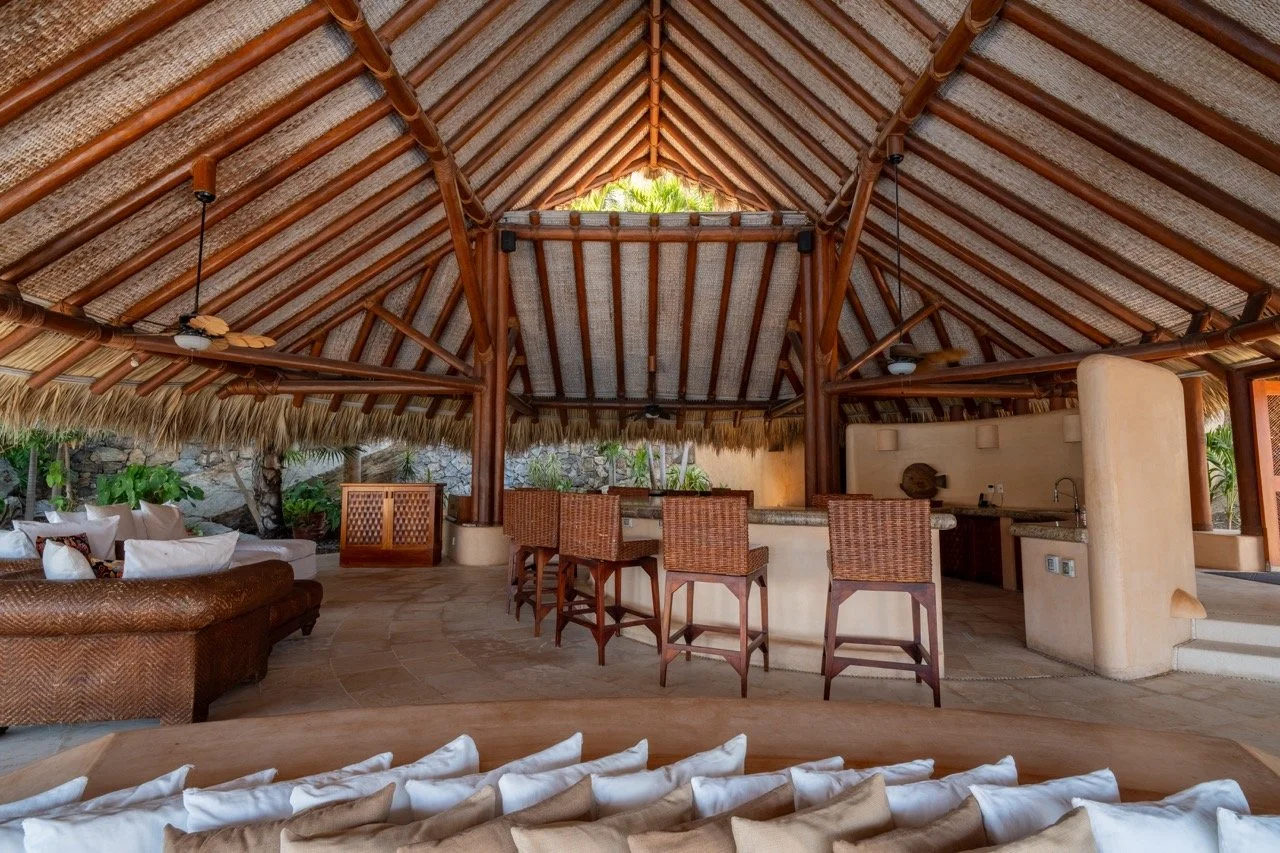 Interior de una cabaña o espacio al aire libre con estructura de madera y techo de paja, bar con asientos de madera, sillón de mimbre, y áreas de descanso con cojines blancos, rodeado de vegetación tropical.