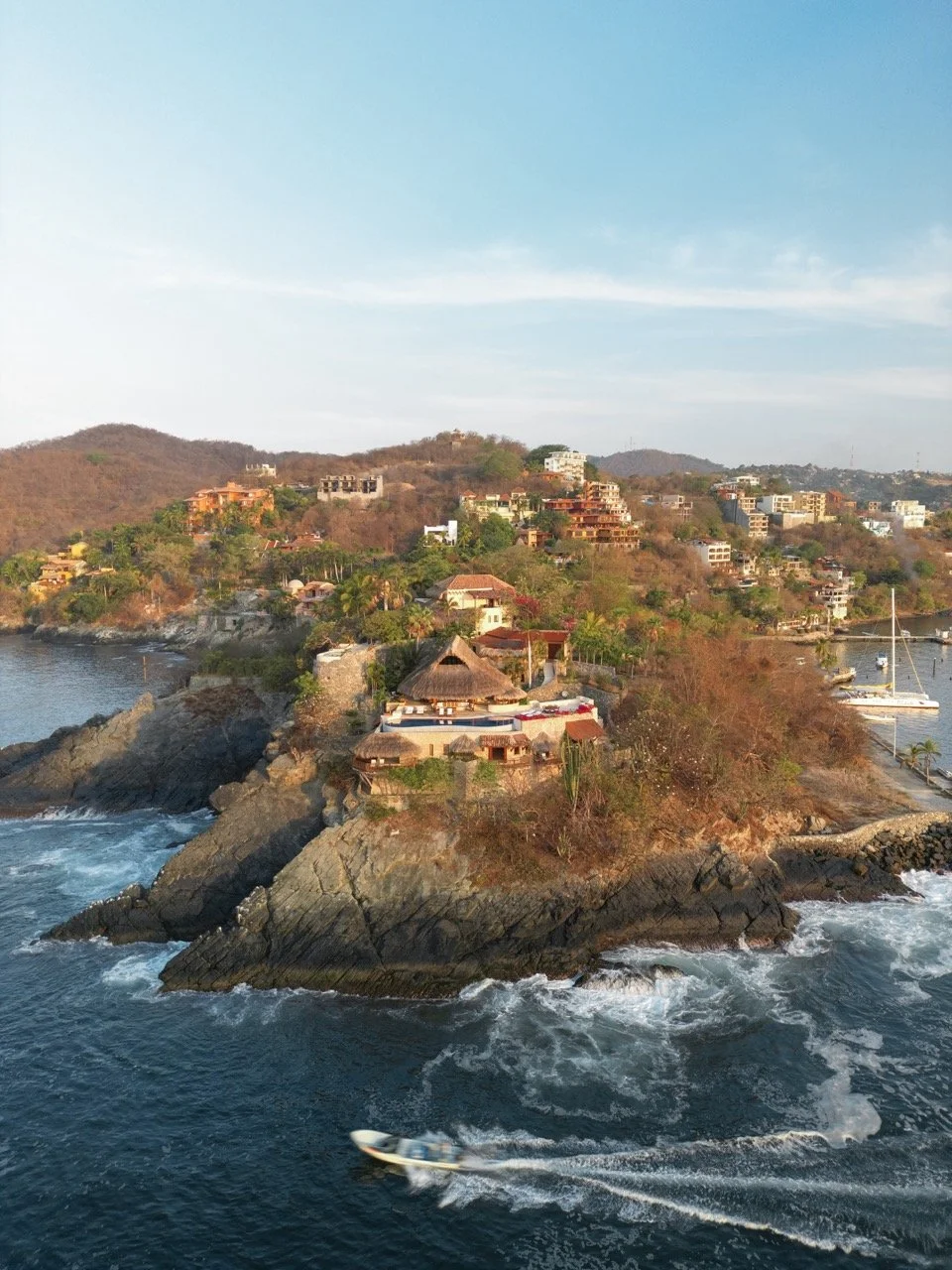 Vista aérea de una zona costera con casas y condominios en las colinas, con barcos en el agua y un bote en movimiento en el mar.