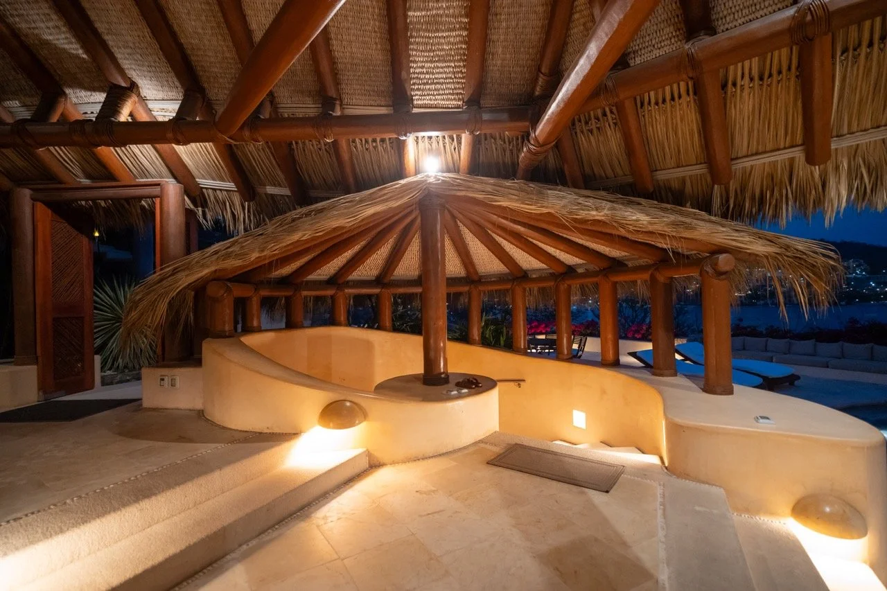 Terraza con piscina cubierta, estructura de madera y techo de paja, iluminada por luces cálidas, con vista nocturna de una ciudad y agua al fondo.