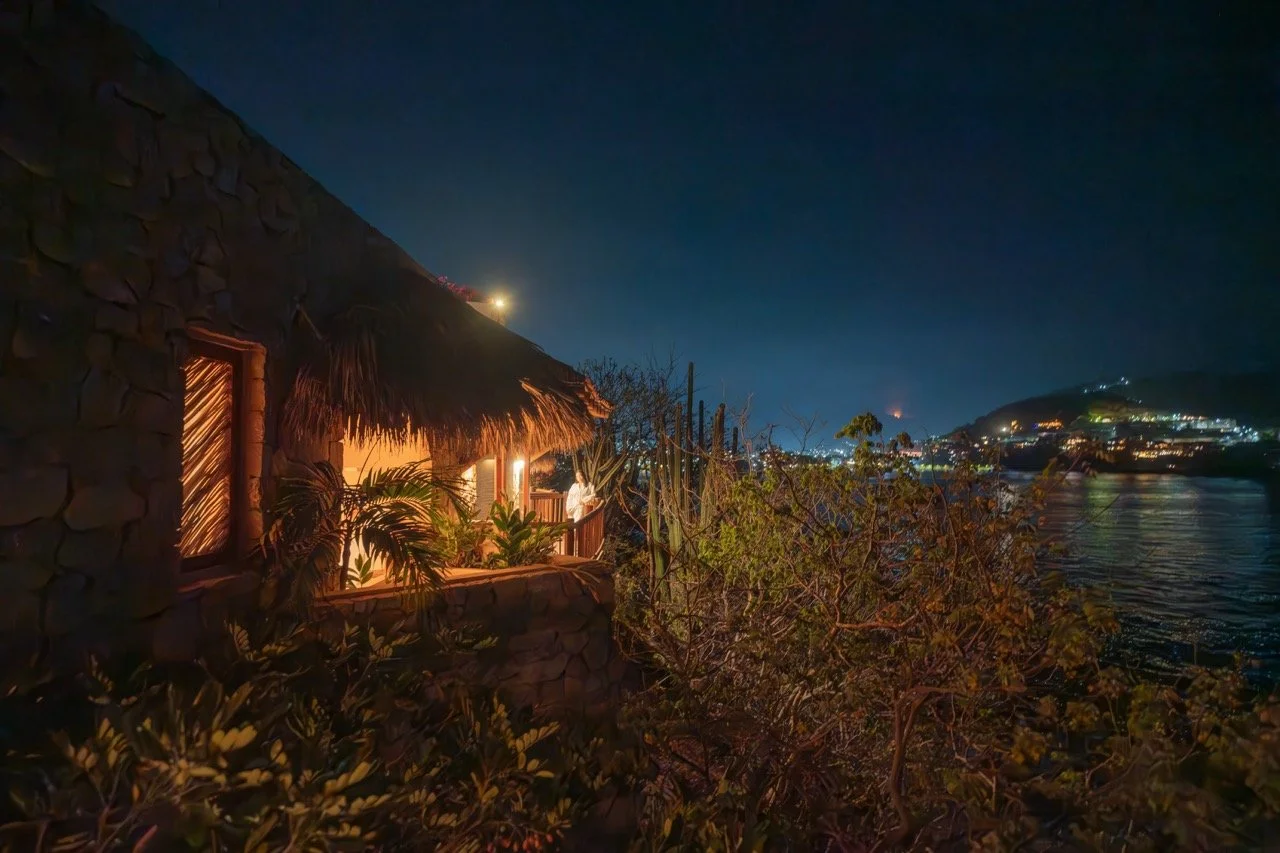 Casa rústica con techo de paja iluminada, junto a un río, con montañas y luces de la ciudad en el fondo durante la noche.