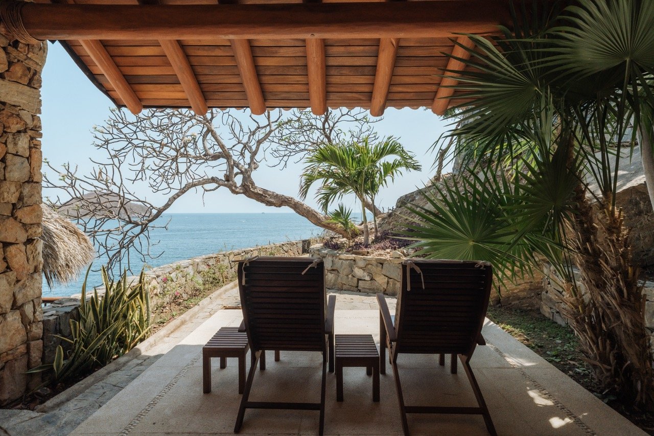 Vista desde una terraza con sillones de madera frente al mar, rodeada de plantas y árboles, con cielo despejado y paisaje costero.