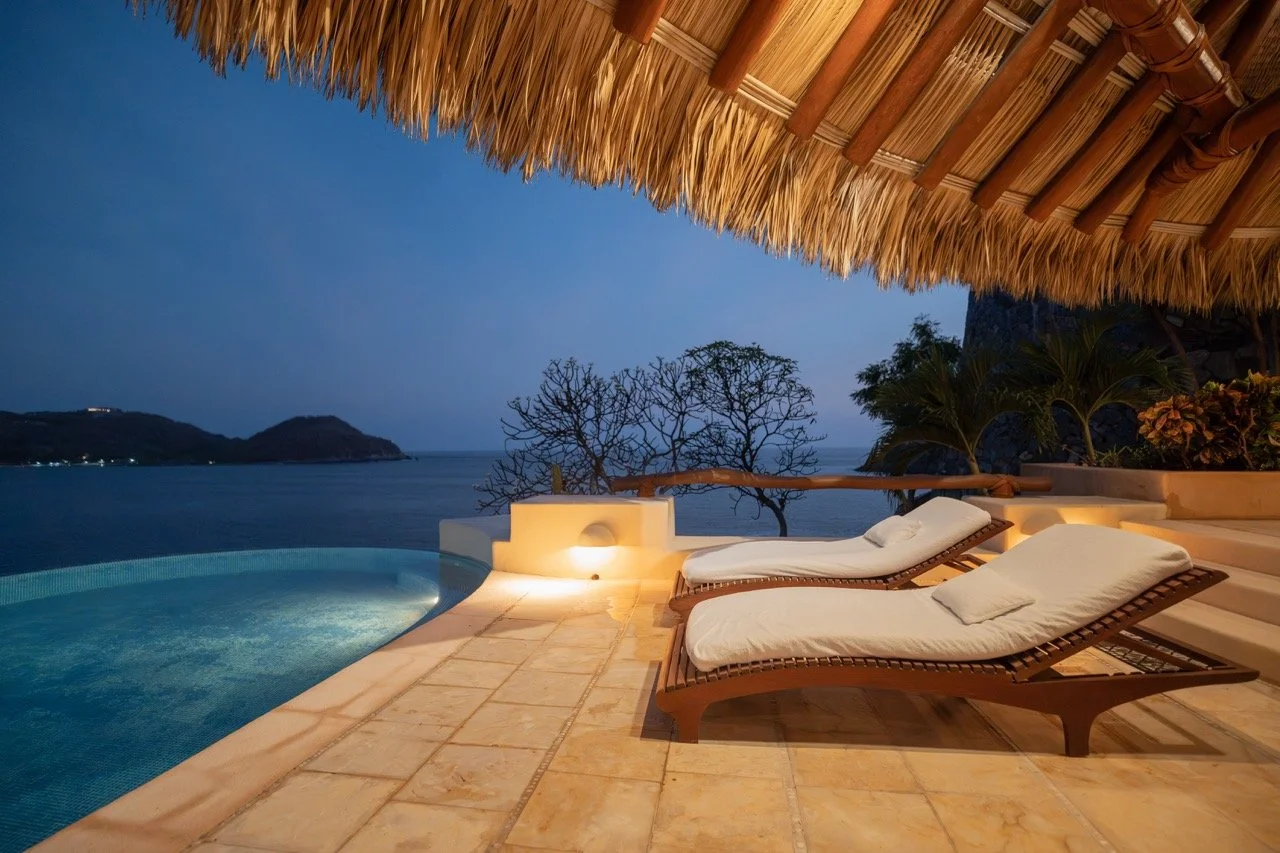 Vista nocturna de una terraza con tumbonas, una piscina y vista al mar y montañas, con techumbre de palma.