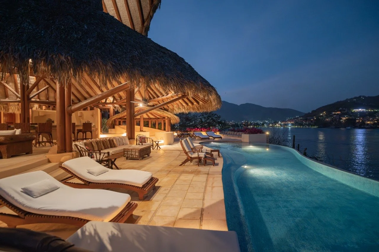 Piscina junto a un río con tumbonas y áreas de descanso en una zona de lujo con cabañas de madera y vista a montañas al atardecer en un entorno paradisíaco.