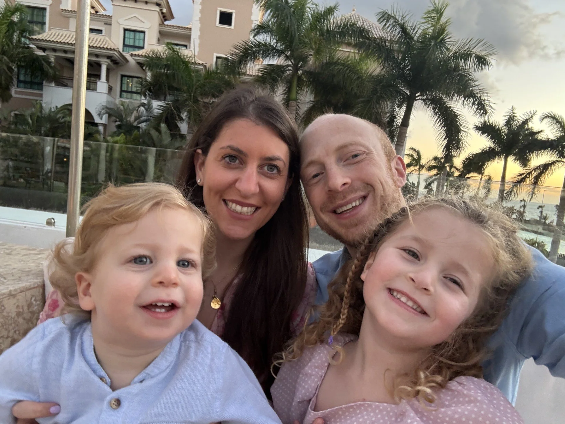 A family taking a selfie at sunset on a patio with palm trees and a large building in the background.