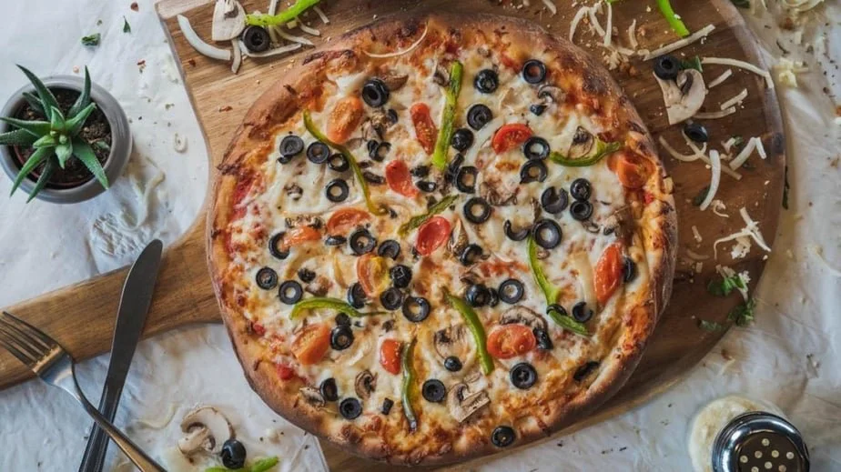 A freshly baked pizza topped with black olives, cherry tomatoes, green peppers, and mushrooms on a wooden pizza board, with some spilled toppings and a small potted succulent nearby.