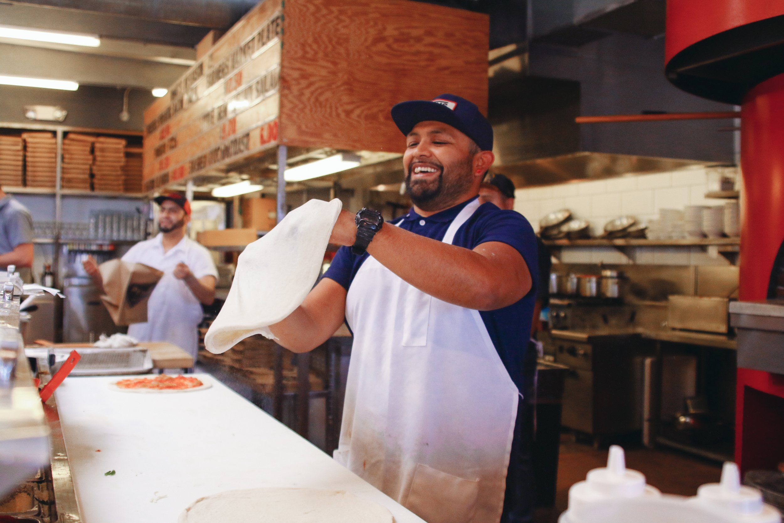 A smiling pizza chef tossing pizza dough in a pizzeria kitchen.