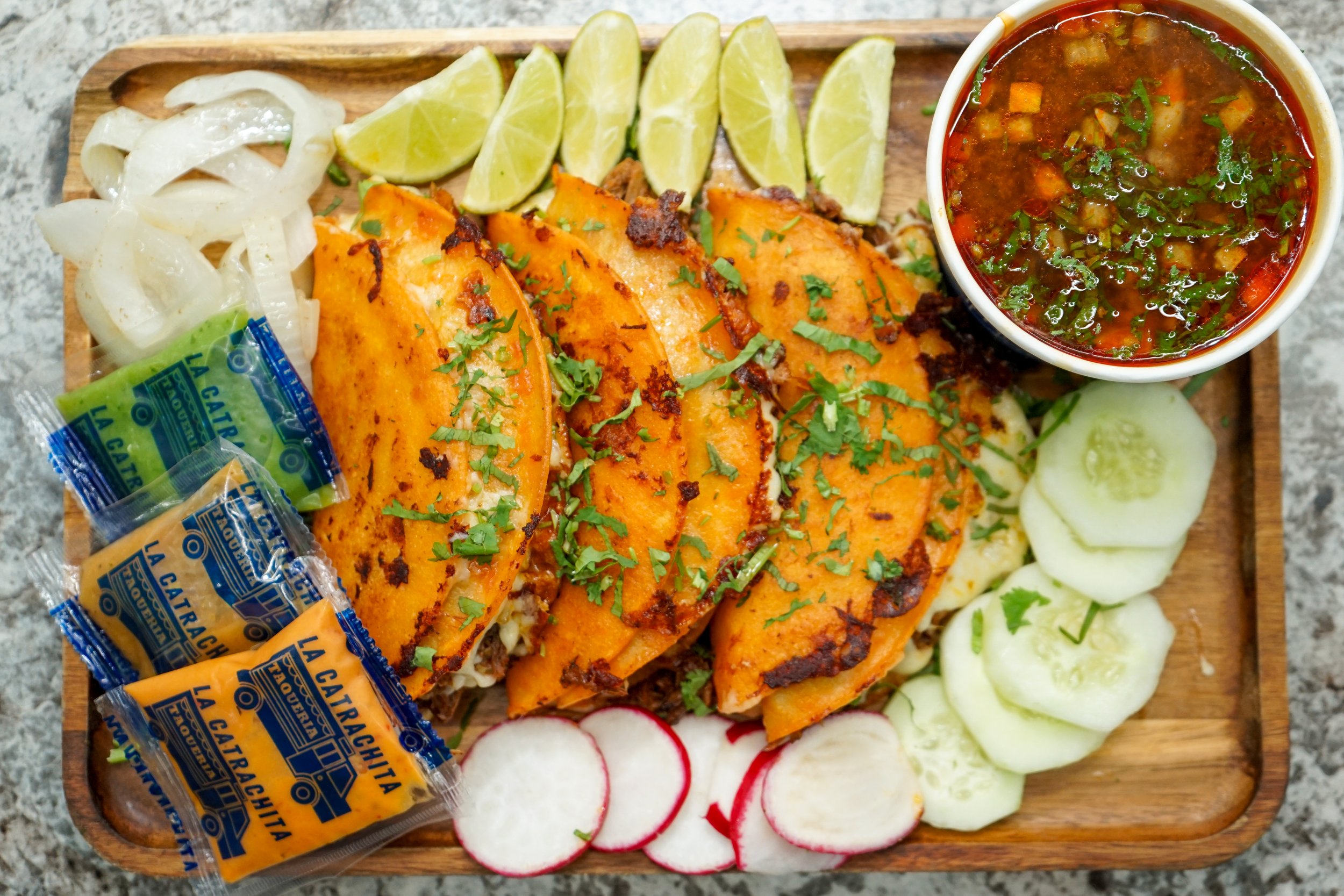 A wooden serving board with grilled fish, lemon wedges, sliced cucumbers, radishes, sliced onions, and a bowl of soup