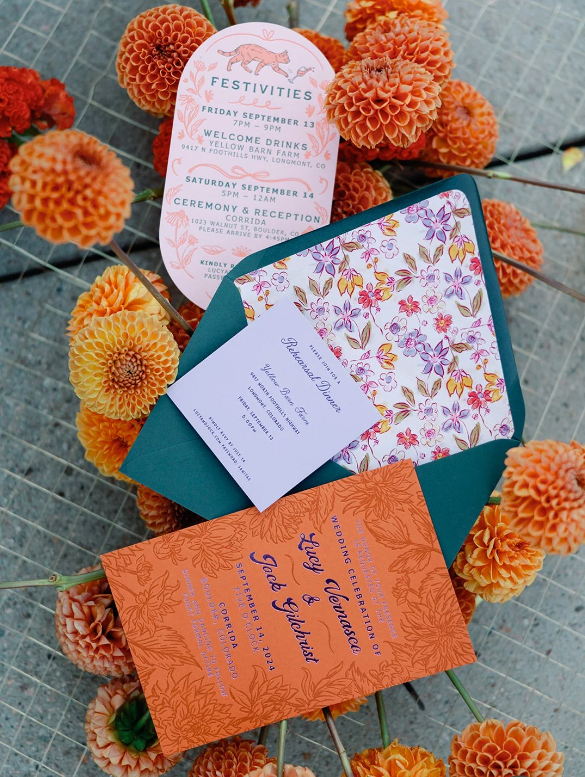 Wedding invitation and event details cards placed among orange, peach, and yellow dahlias on a wire mesh surface.